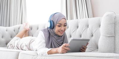 Relaxed and happy asian Muslim woman with hijab laying on sofa, listening music on headphones and using digital tablet. photo