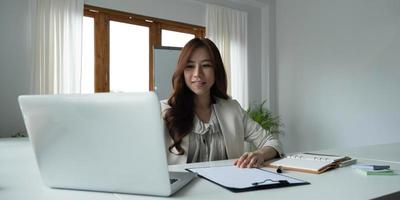 hermosa joven asiática sentada en una cafetería usando una laptop. feliz joven mujer de negocios sentada a la mesa en un café con una computadora con pestaña. foto