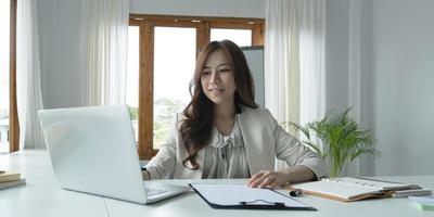 hermosa joven asiática sentada en una cafetería usando una laptop. feliz joven mujer de negocios sentada a la mesa en un café con una computadora con pestaña. foto