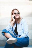 Photo of a cheerful smiling cute young student girl wearing sunglasses outdoors using mobile phone chatting listening music with earphones.