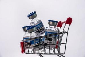 viales de vidrio para la vacuna covid-19 en carrito de compras sobre fondo blanco. grupo de botellas de vacuna contra el coronavirus en la cesta. foto