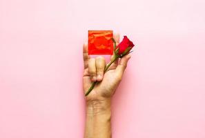 Hand posing love symbol with rose and condom package for minimal safe sex, world sexual health and aids day concept. photo