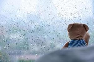 Lonely Teddy Bear sitting on bed and looking out at the window in rainy day. photo