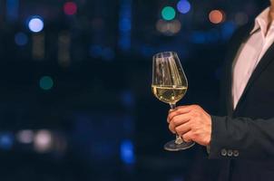 Businessman wearing navy blue color suit stands at the rooftop bar toasting a glass of white wine with dark background of city bokeh lights. photo