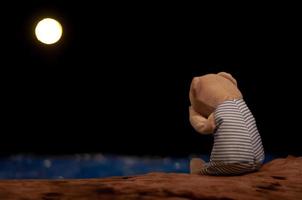 Teddy bear crying and sitting alone facing to the blue sea and moon. photo