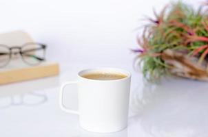 una taza de café en la mesa con planta de aire tillandsia, espectáculos y libro sobre fondo blanco. foto