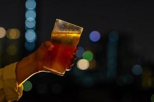 mano con un vaso de cerveza brindando por concepto de celebración y fiesta aislado en el fondo oscuro de la noche con coloridas luces de ciudad en el bar de la azotea. foto