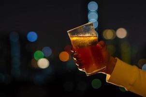 Hand with glass of beer toasting for celebration and party concept isolated on dark night background with colorful city bokeh lights on rooftop bar. photo