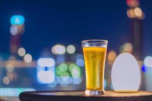A glass of beer puts on table at rooftop bar with colorful city bokeh light background.