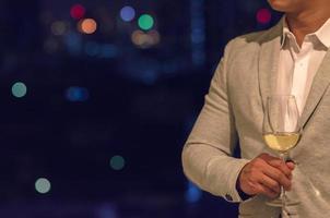 un hombre de negocios con traje de color gris se encuentra en el bar de la azotea sosteniendo una copa de vino blanco con un fondo oscuro de luces de bokeh de la ciudad.