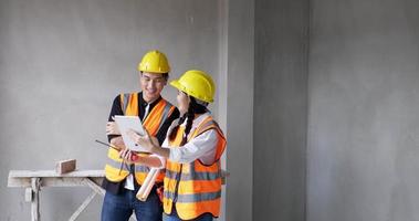 From work at a building site indoors, a young female and male foreman wearing orange vests and safety helmets are video calling on their tablets.