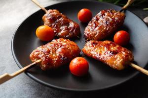 Close-up grill skewered milk pork in a black dish with tomatoes with garlic and spring onion on a gray concrete background. Thai street food photo