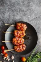 Grill skewered milk pork in a black dish with tomatoes with garlic and spring onion on a gray concrete background. Thai street food.Top view and copy space photo