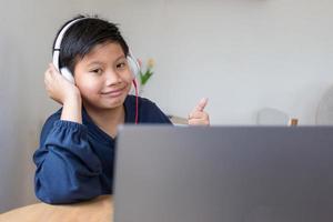 chico asiático chico lindo usa auriculares que muestran los pulgares hacia arriba y sonríe mientras mira a la cámara después de estudiar en línea con una computadora portátil en casa. foto