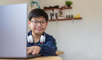 un estudiante asiático inteligente que usa anteojos con auriculares mirando la cámara y sonriendo, usa una computadora portátil para usar Internet en las redes sociales sentado en casa. foto