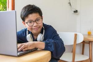 un estudiante asiático inteligente que usa anteojos con auriculares mirando la cámara y sonriendo, usa una computadora portátil para usar Internet en las redes sociales sentado en casa. foto