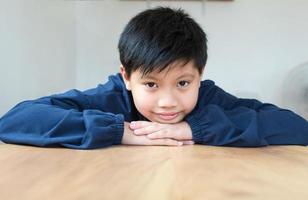 Cute Asian boy with white skin smiling looking in camera happily resting chins on hands-on wooden table. Close-up portrait of a cute child photo