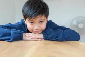 Cute Asian boy with white skin smiling looking in camera happily resting chins on hands-on wooden table. Close-up portrait of a cute child photo