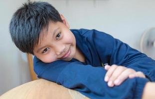 Cute Asian boy with white skin smiling looking in camera happily resting chins on hands-on wooden table. Close-up portrait of a cute child photo