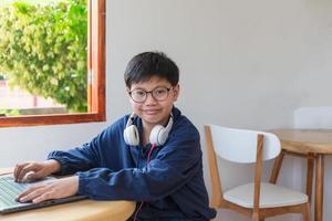 Smart Asian student boy wearing glasses with headphones looking at the camera and smiling, he using a computer laptop to use the internet on social networks sitting at home. photo