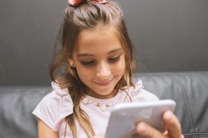 Beautiful little girl playing game or watching video on smartphone mobile. Girl watching cartoons or browsing internet, copy space. Portrait of little girl using smartphone while sitting. photo