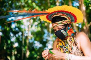 indio de la tribu pataxo con tocado de plumas y máscara protectora. mujer indígena de brasil haciendo artesanías foto