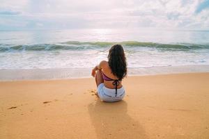 silueta de mujer joven en la playa. mujer latinoamericana sentada en la arena de la playa mirando al cielo en un hermoso día de verano foto