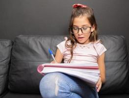 Little girl writing in copybook. Beautiful elementary schoolgirl studying in sofa. photo