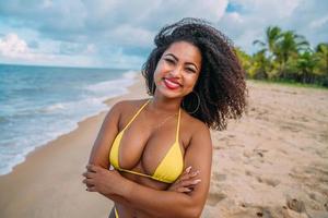 hermosa mujer latinoamericana en bikini en la playa. mujer joven disfrutando de sus vacaciones de verano en un día soleado, sonriendo, con los brazos cruzados y mirando la cámara foto