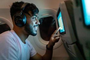 Passenger in airplane touching LCD entertainment screen. Latin american man in plane cabin using smart device listening to music on headphones. photo