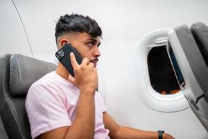 un joven latinoamericano está hablando por teléfono celular mientras está sentado en el avión cerca de la ventana. foto