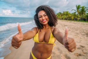 hermosa mujer latinoamericana en bikini en la playa. mujer joven disfrutando de sus vacaciones de verano en un día soleado, sonriendo, con los pulgares hacia arriba y mirando la cámara foto