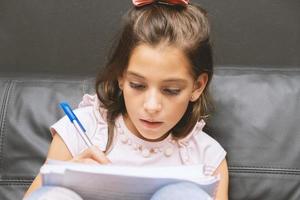 niña escribiendo en cuaderno. hermosa colegiala de primaria estudiando en el sofá. foto