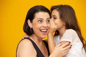 madre e hija en secreto. niño compartiendo un secreto con su madre. mamá pequeña hija aislada en un retrato de estudio de fondo amarillo. día de la madre amor familia paternidad infancia concepto foto