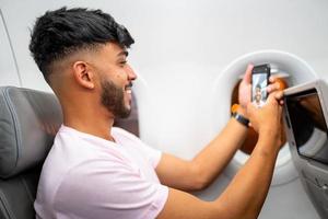 joven latinoamericano sonriendo haciendo una videollamada en su celular, sentado en el avión cerca de la ventana. foto