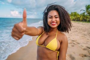 beautiful Latin American woman in bikini on the beach. Young woman enjoying her summer vacation on a sunny day, smiling, with thumbs up and looking at the camera photo