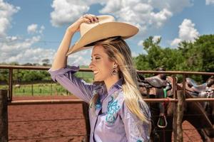 hermosa vaquera rubia con sombrero de pie cerca del fondo del rancho de caballos foto