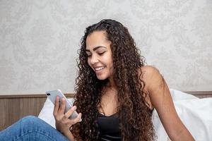 Technology, free time, communication concept. Cheerful Afro American, dressed on bed, in hotel or home, uses mobile phone for online chatting, networking, receives text message photo