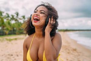 vacaciones de verano, tecnología y concepto de internet. mujer latinoamericana escuchando música con auriculares y tomando el sol en la playa foto