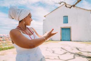 mujer brasileña feliz vestida con traje tradicional bahiano que muestra la iglesia con la palma de su mano, mira a la cámara, con el centro histórico de porto seguro al fondo foto