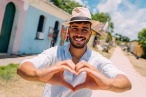 Tourist making selfie in the historic center of Porto Seguro. Latin American man in a hat, making a heart with his hands, smiling at the camera photo