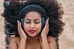 summer holidays, technology and internet concept. latin american woman listening music with headphone and sunning on the beach photo