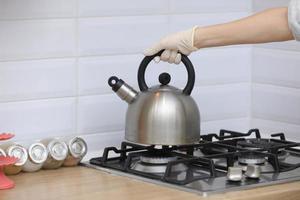 la mujer pone una tetera en la cocina con guantes en la estufa de gas. tetera silbante en la estufa en la mano. enfoque selectivo foto