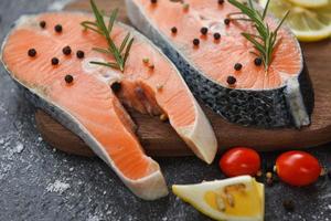 Raw salmon fish fillet and ice for cooked steak seafood - Fresh salmon steak with herbs and spices lemon rosemary tomato on wooden cutting board background photo