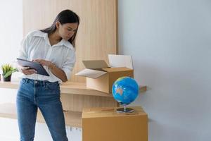 Asian woman checks parcel boxes and checks orders from laptop She owns an online store. Online Selling and Shopping Ideas photo