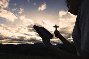 silueta de mujer rezando con cruz en el fondo del amanecer de la naturaleza, crucifijo, símbolo de fe. oración de crisis de vida cristiana a dios. foto