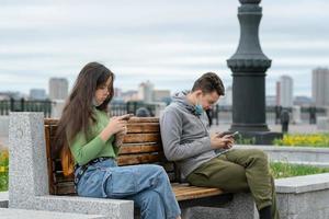 social distance, a guy and a girl are sitting on a bench at a distance from each other with masks on their faces and with phones in their hands photo