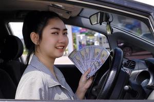 Retrato cerrado de una atractiva mujer asiática feliz y sonriente que sostiene billetes de dólar en efectivo sentados dentro de su auto en la gasolinera foto