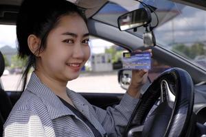 Happy beautiful Asian woman sitting inside her car showing credit card  payment at a gas station photo