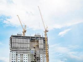 dos grúas de construcción están trabajando en la construcción de edificios altos, levantando y colocando cosas. el sitio de construcción está cubierto con mantas. en el cielo despejado edificio de la ciudad en línea recta foto